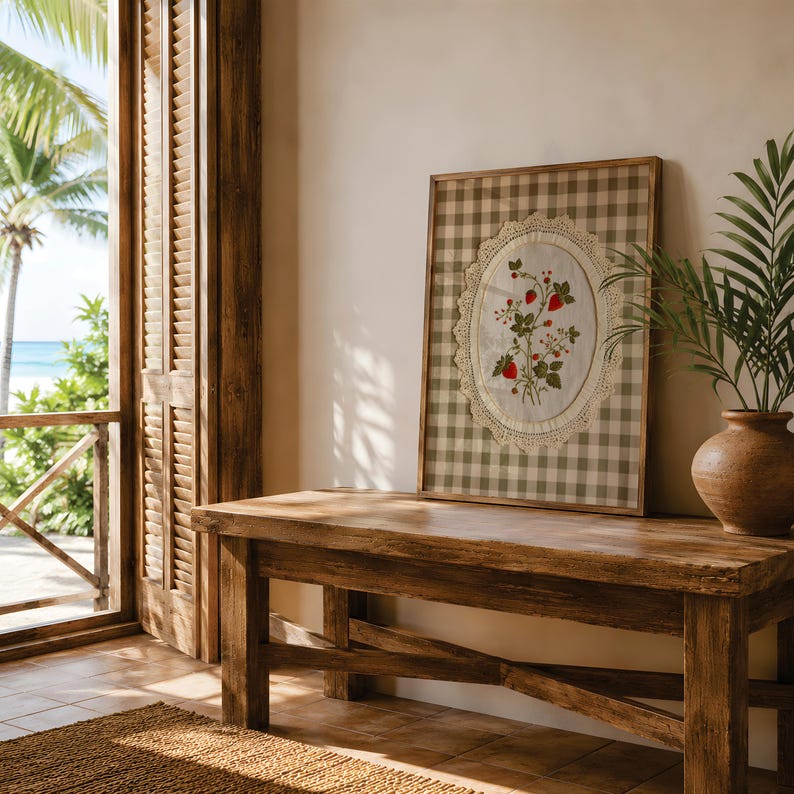Op de afbeelding: Een rechthoekig ingelijst kunstwerk met een beige en witte gingham-achtergrond. In het midden bevindt zich een ovaal stoffen inzetstuk met een botanische illustratie van rode aardbeien en groene bladeren, omrand door een cr&egrave;mekleurige gehaakte kanten rand.