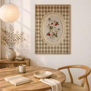 Op de afbeelding: Een rechthoekig wandkunstwerk met een beige en witte gingham-achtergrond en een centraal ovaal stoffen paneel. Het paneel is omlijst met cr&egrave;mekleurig kant en toont een geborduurd botanisch ontwerp van groene bladeren en rode aardbeien.