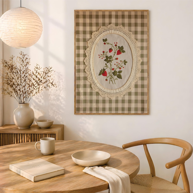 Op de afbeelding: Een rechthoekig wandkunstwerk met een beige en witte gingham-achtergrond en een centraal ovaal stoffen paneel. Het paneel is omlijst met cr&egrave;mekleurig kant en toont een geborduurd botanisch ontwerp van groene bladeren en rode aardbeien.
