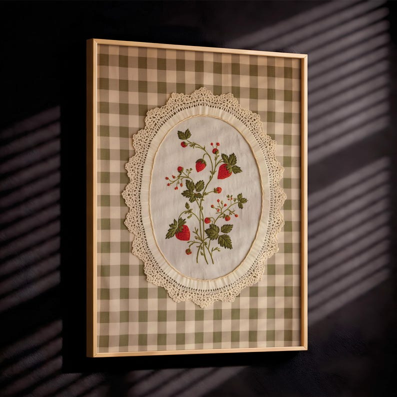 Op de afbeelding: Een ingelijst wandkunstwerk met een wit stoffen ovaal gecentreerd op een saliegroene en witte gingham-achtergrond. Het ovaal is omrand met cr&egrave;mekleurig kant en toont een geborduurde botanische illustratie van rode aardbeien en groene bladeren.
