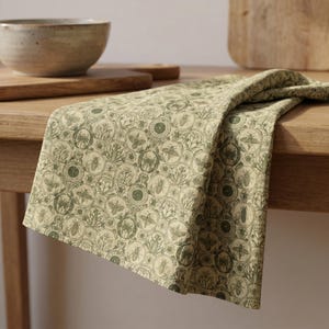 May include: A folded green and beige patterned kitchen towel with a repeating design of bees and botanical elements. The towel is draped over a wooden table, with a bowl and cutting board in the background.