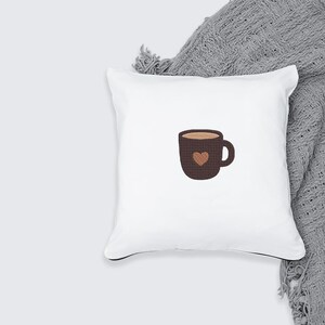 K&ouml;nnte beinhalten: Wei&szlig;es quadratisches Kissen mit einem braunen Kaffeetassen-Design. Die Tasse hat eine Herzform und ist mit einer hellbraunen Fl&uuml;ssigkeit gef&uuml;llt. Das Kissen liegt auf einer grauen Decke.