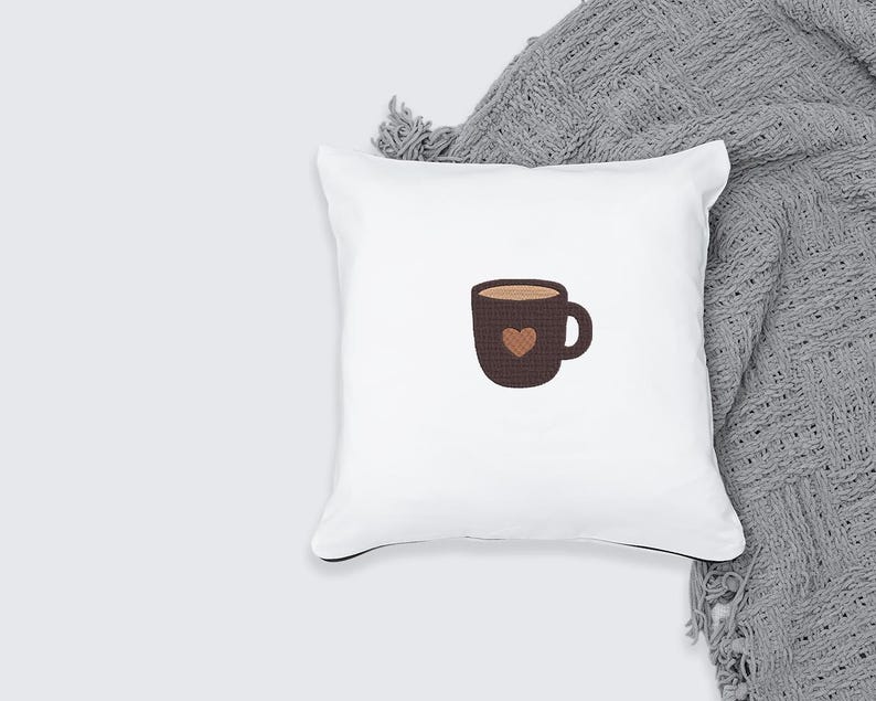 K&ouml;nnte beinhalten: Wei&szlig;es quadratisches Kissen mit einem braunen Kaffeetassen-Design. Die Tasse hat eine Herzform und ist mit einer hellbraunen Fl&uuml;ssigkeit gef&uuml;llt. Das Kissen liegt auf einer grauen Decke.