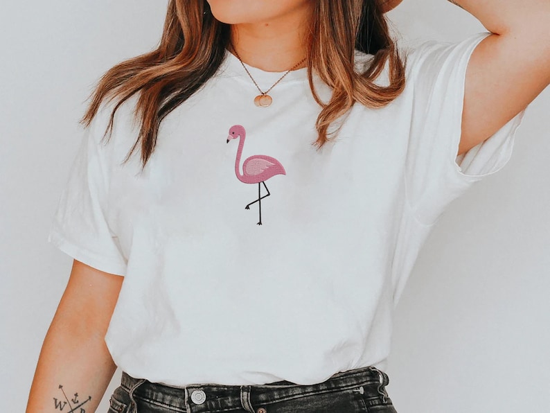 May include: White t-shirt featuring a pink flamingo design. The flamingo is embroidered in shades of pink with black legs and beak. The shirt is a casual style, suitable for everyday wear.
