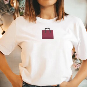 May include: White t-shirt featuring a burgundy shopping bag graphic with black handles. The t-shirt is worn with blue jeans. The person has short brown hair.