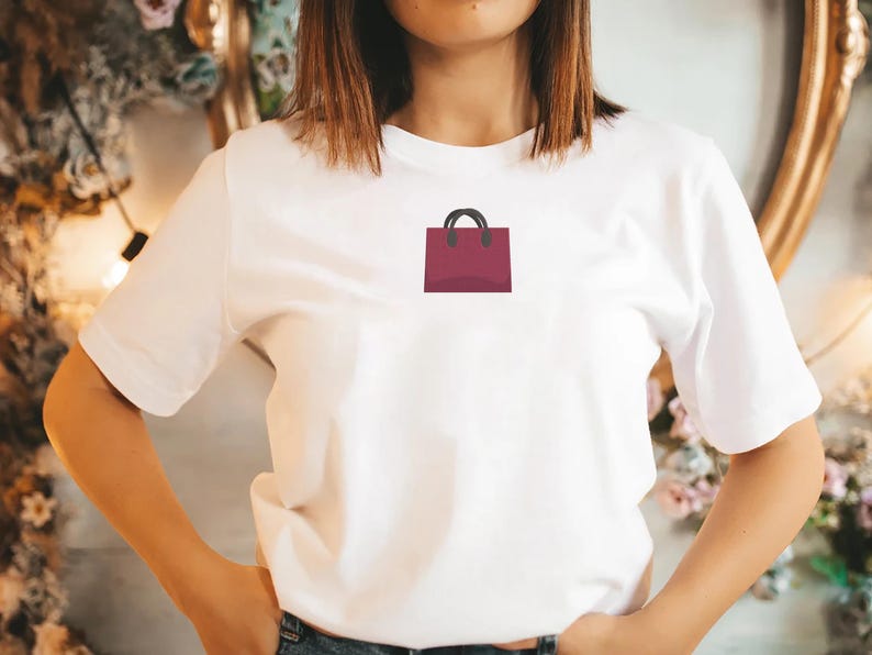 May include: White t-shirt featuring a burgundy shopping bag graphic with black handles. The t-shirt is worn with blue jeans. The person has short brown hair.