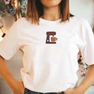 K&ouml;nnte beinhalten: Wei&szlig;es T-Shirt mit einem kleinen, gestickten Kaffeemaschinen- und Tassen-Design in Braunt&ouml;nen. Ein l&auml;ssiges Kleidungsst&uuml;ck f&uuml;r den Alltag, ideal f&uuml;r Kaffeeliebhaber.