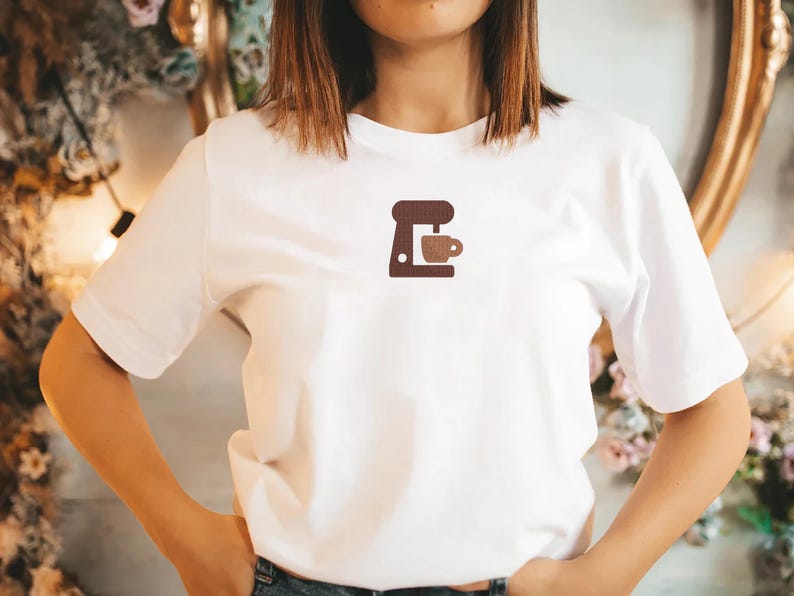 K&ouml;nnte beinhalten: Wei&szlig;es T-Shirt mit einem kleinen, gestickten Kaffeemaschinen- und Tassen-Design in Braunt&ouml;nen. Ein l&auml;ssiges Kleidungsst&uuml;ck f&uuml;r den Alltag, ideal f&uuml;r Kaffeeliebhaber.