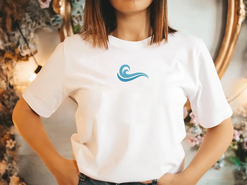 May include: White t-shirt with a small, blue wave design on the chest. The short-sleeved shirt is made of a soft material and is suitable for casual wear. The wave graphic is a simple, stylized design.