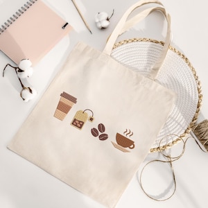 K&ouml;nnte beinhalten: Cremefarbene Tragetasche mit gestickten Kaffee- und Tee-Motiven. Das Design umfasst eine Kaffeetasse, einen Teebeutel, Kaffeebohnen und einen Coffee-to-go-Becher. Die Tasche hat zwei Henkel und liegt auf einer wei&szlig;en Oberfl&auml;che mit Notizbuch und Bleistift.