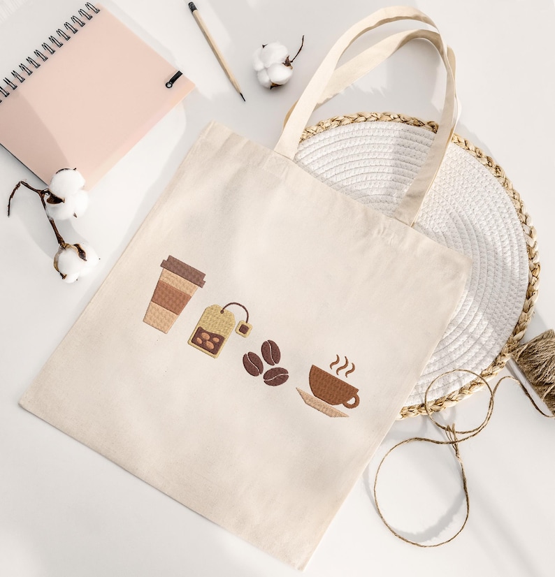 K&ouml;nnte beinhalten: Cremefarbene Tragetasche mit gestickten Kaffee- und Tee-Motiven. Das Design umfasst eine Kaffeetasse, einen Teebeutel, Kaffeebohnen und einen Coffee-to-go-Becher. Die Tasche hat zwei Henkel und liegt auf einer wei&szlig;en Oberfl&auml;che mit Notizbuch und Bleistift.