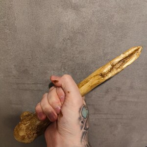 Può includere: Una mano tiene un lungo bastone di legno marrone con un'estremità arrotondata. Il bastone è liscio e leggermente curvo. La mano è tatuata con un disegno nero e verde.