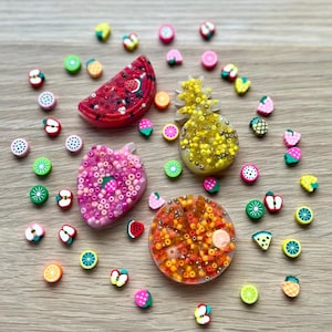 May include: A selection of colourful fruit-shaped resin pieces and fruit-themed beads. The pieces include a watermelon slice, pineapple, strawberry, and orange slice, all filled with small beads. Various fruit-shaped beads are scattered around.