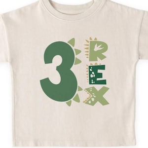 May include: Cream-colored toddler t-shirt with the number "3" and the word "REX" in green, dinosaur-themed font. The "3" is a solid green, while the letters of "REX" have a textured, scaly design. The shirt is made of a soft material.