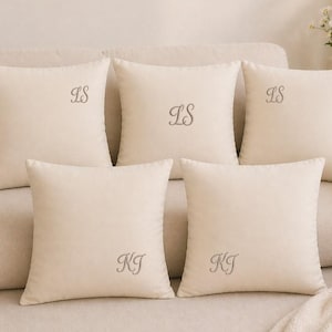 May include: Five cream-coloured square throw cushions with embroidered initials on a beige sofa. The cushions have the initials "LS" and "KJ" embroidered in a grey thread. The sofa is covered in a neutral-toned fabric.