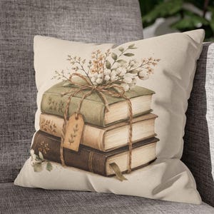 May include: A decorative cushion with a print of a stack of old books tied with twine and decorated with flowers. The books are in shades of green, brown, and beige, creating a cosy, literary theme. The cushion is on a grey sofa.