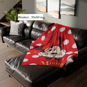 May include: A red blanket with white polka dots and a Minnie Mouse graphic. The blanket measures 50 x 60 inches and features the word "Minnie" in gold lettering. The blanket is draped over a black leather couch.