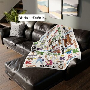 May include: A white blanket with colorful cartoon characters and the name "Kieran" printed repeatedly. The blanket, measuring 50x60 inches, features images of a rocket, a horse, and other characters. It is draped over a dark brown leather couch.