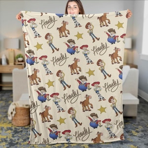 May include: A beige blanket with a repeating pattern of cartoon characters, including a cowboy, a horse, and a sheriff. The word "Howdy" is written in a cursive font. Yellow stars are also included in the design.