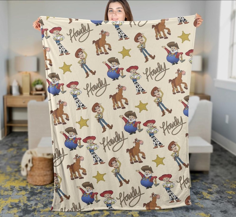 May include: A beige blanket with a repeating pattern of cartoon characters, including a cowboy, a horse, and a sheriff. The word "Howdy" is written in a cursive font. Yellow stars are also included in the design.