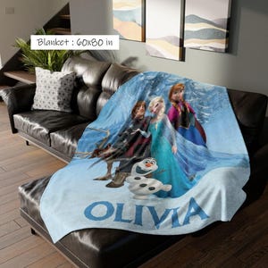 May include: A light blue blanket, 60x80 inches, featuring characters from the movie Frozen. The blanket displays Elsa, Anna, Kristoff, and Olaf, with the name "Olivia" in large blue letters.