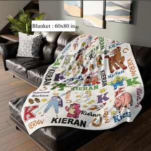 May include: A white blanket with a colorful cartoon design featuring characters and the name "Kieran". The blanket measures 60x80 inches and is draped over a black leather couch. The blanket has a variety of cartoon characters.