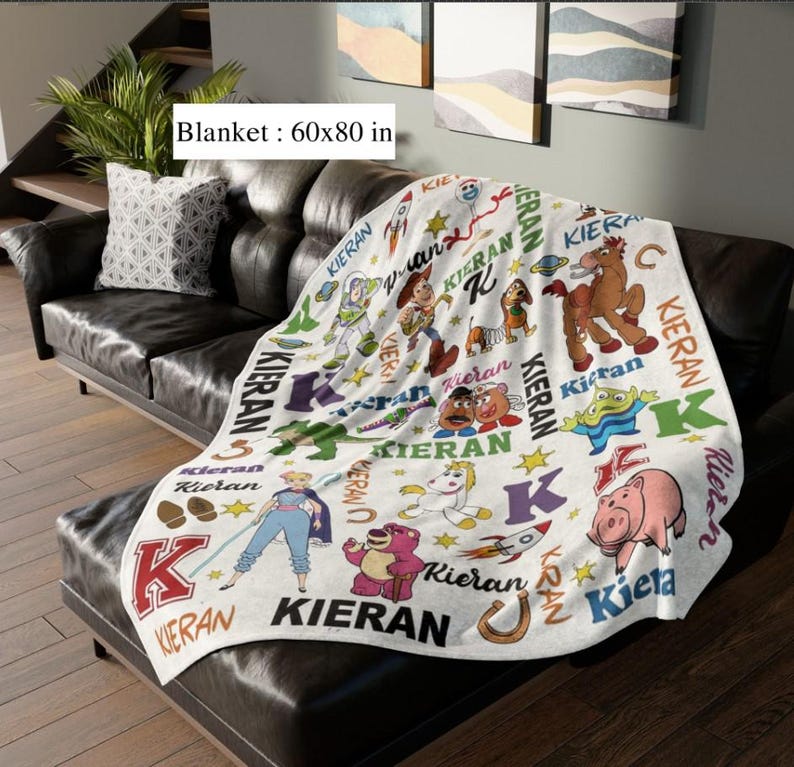 May include: A white blanket with a colorful cartoon design featuring characters and the name "Kieran". The blanket measures 60x80 inches and is draped over a black leather couch. The blanket has a variety of cartoon characters.