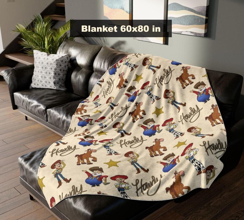 May include: A beige blanket featuring a repeating pattern of cartoon cowboys, horses, and stars. The blanket measures 152x203 cm. The word "Howdy" is written in a cursive font. The blanket is draped over a black leather sofa.
