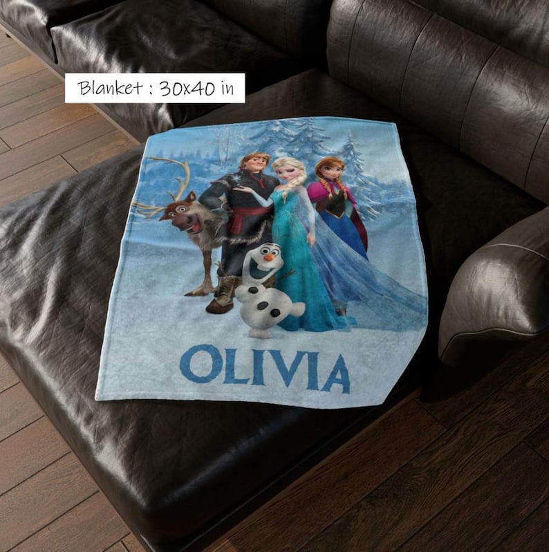 May include: A light blue blanket, 30 x 40 inches, featuring characters from the movie "Frozen." The blanket displays Elsa, Anna, Kristoff, Sven, and Olaf in a snowy scene. The name "OLIVIA" is printed in blue.