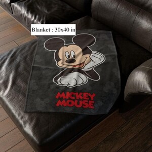 May include: A black blanket with a Mickey Mouse graphic. The blanket, measuring 76 x 102 cm, has the words "Mickey Mouse" in red. The blanket is placed on a black leather sofa.