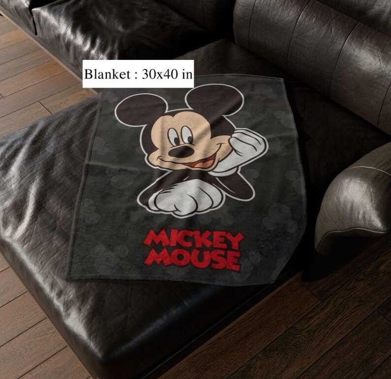 May include: A black blanket with a Mickey Mouse graphic. The blanket, measuring 76 x 102 cm, has the words "Mickey Mouse" in red. The blanket is placed on a black leather sofa.