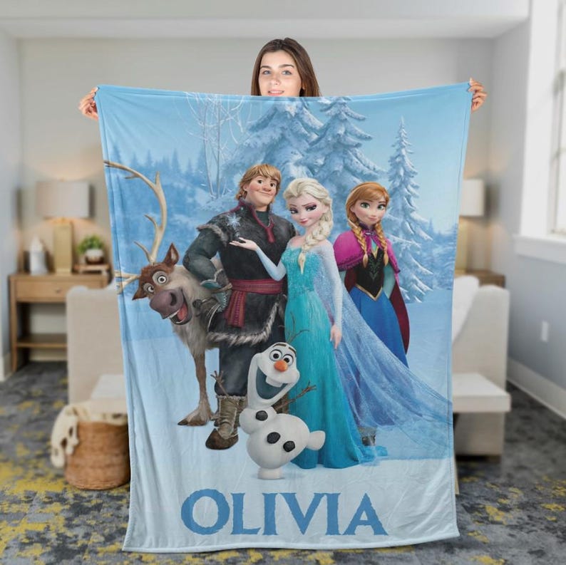 May include: A light blue fleece blanket featuring characters from the movie Frozen, including Elsa, Anna, Kristoff, Sven, and Olaf, set against a snowy forest backdrop. The name "OLIVIA" is printed in blue at the bottom.