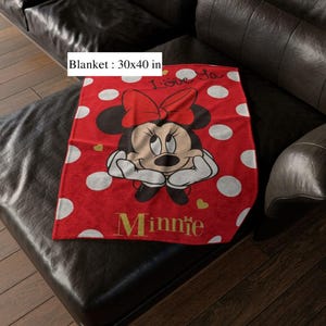 May include: A red blanket with white polka dots and a Minnie Mouse graphic. The blanket, measuring 30 x 40 inches, features Minnie Mouse with a red bow and the word "Minnie" in gold lettering. The blanket also has the words "I love ya" in white.