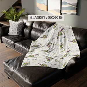 May include: A white blanket with a cartoon character design, measuring 50 x 60 inches, is draped over a black leather couch. The blanket features a repeating pattern of a green, purple, and white cartoon character, space ships, and planets.