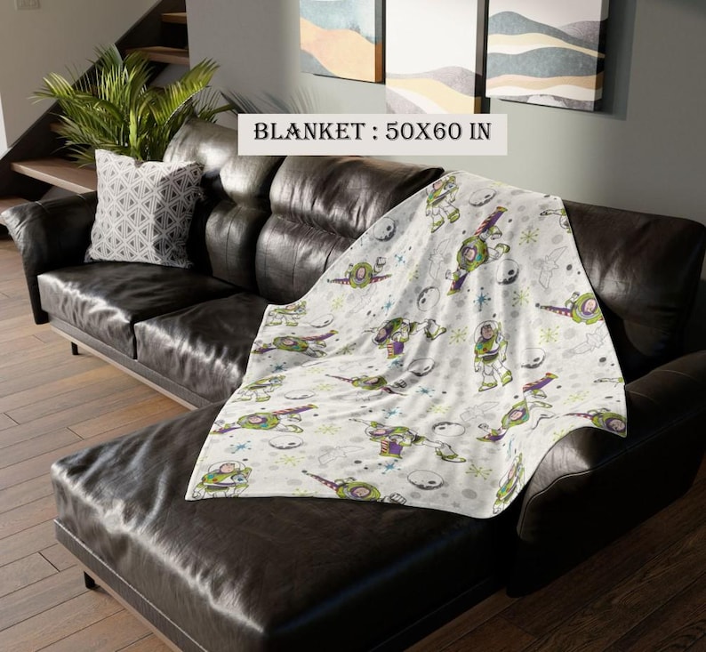 May include: A white blanket with a cartoon character design, measuring 50 x 60 inches, is draped over a black leather couch. The blanket features a repeating pattern of a green, purple, and white cartoon character, space ships, and planets.