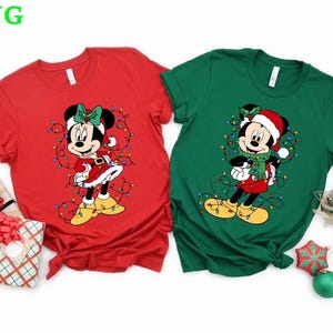 May include: Two short-sleeved Christmas-themed t-shirts. One is red with a Minnie Mouse graphic, the other is green with a Mickey Mouse graphic. Both characters are dressed in holiday attire and surrounded by string lights. Festive apparel.