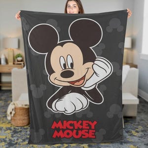 May include: A dark grey blanket with a large illustration of Mickey Mouse. The cartoon character is smiling with his right hand raised. The words "Mickey Mouse" are printed in red at the bottom of the blanket. Mickey Mouse silhouettes are in the background.