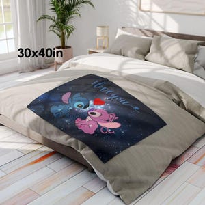 May include: A square throw blanket, 30 x 40 inches, featuring a blue and pink cartoon character design with the words "I love you" and a red heart. The blanket is placed on a bed with neutral-colored bedding.