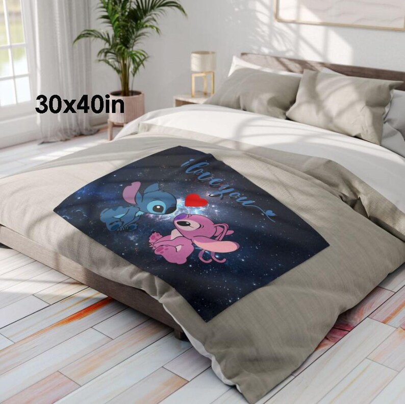 May include: A square throw blanket, 30 x 40 inches, featuring a blue and pink cartoon character design with the words "I love you" and a red heart. The blanket is placed on a bed with neutral-colored bedding.