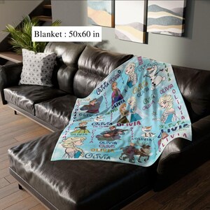 May include: A light blue blanket, measuring approximately 127 x 152 cm, featuring cartoon characters and the name "Olivia". The blanket is draped over a dark brown leather couch, with a decorative pillow.