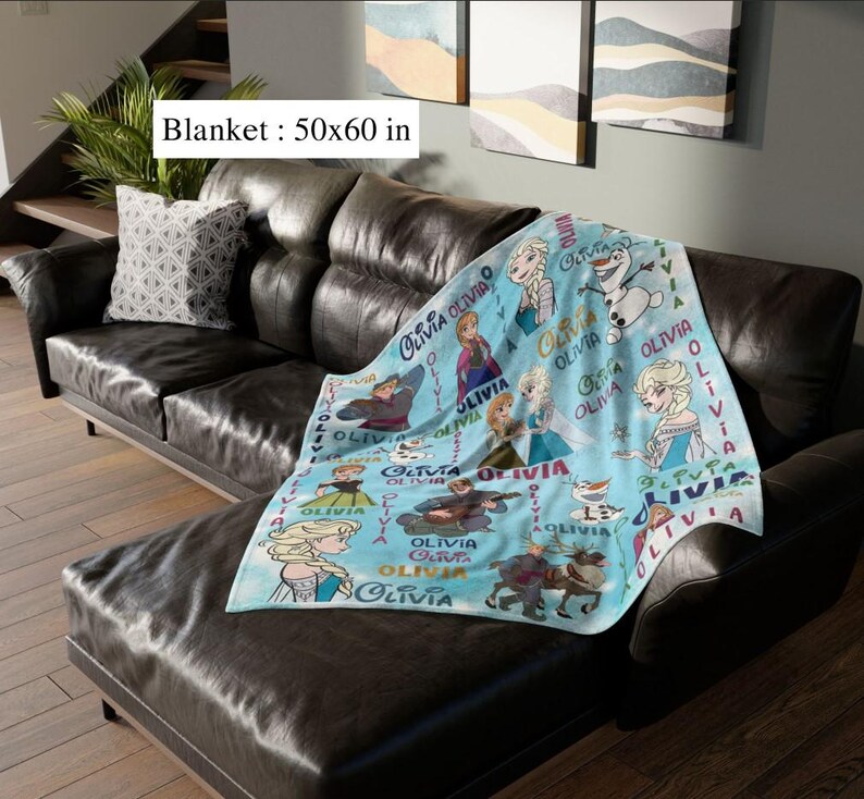 May include: A light blue blanket, measuring approximately 127 x 152 cm, featuring cartoon characters and the name "Olivia". The blanket is draped over a dark brown leather couch, with a decorative pillow.