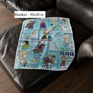 May include: A light blue blanket, measuring 76 x 102 cm, featuring illustrations of characters from a popular animated film. The blanket has the name "Olivia" printed multiple times. The blanket is on a dark leather couch.