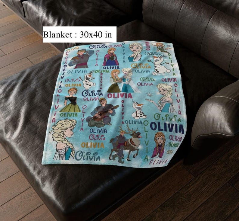 May include: A light blue blanket, measuring 76 x 102 cm, featuring illustrations of characters from a popular animated film. The blanket has the name "Olivia" printed multiple times. The blanket is on a dark leather couch.