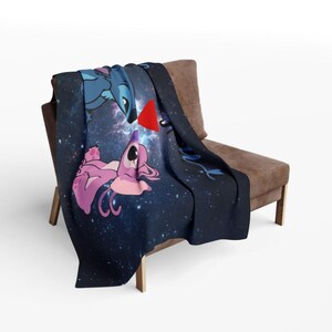 May include: A navy blue throw blanket featuring a space-themed design with cartoon characters. The blanket showcases a blue alien and a pink alien with a red heart between them. The blanket is draped over a brown chair.