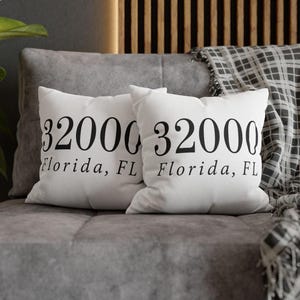 May include: Two square white throw pillows featuring the black printed text 32000 and Florida, FL. The pillows have a smooth fabric texture and are displayed side by side, showcasing the identical numerical and geographical location design.