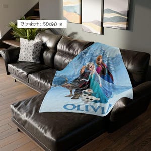May include: A blue blanket featuring characters from the movie Frozen, with the name "OLIVIA" printed in blue. The blanket measures 50 x 60 inches and is draped over a black leather couch. The blanket's design includes Elsa, Anna, Kristoff, Sven, and Olaf.