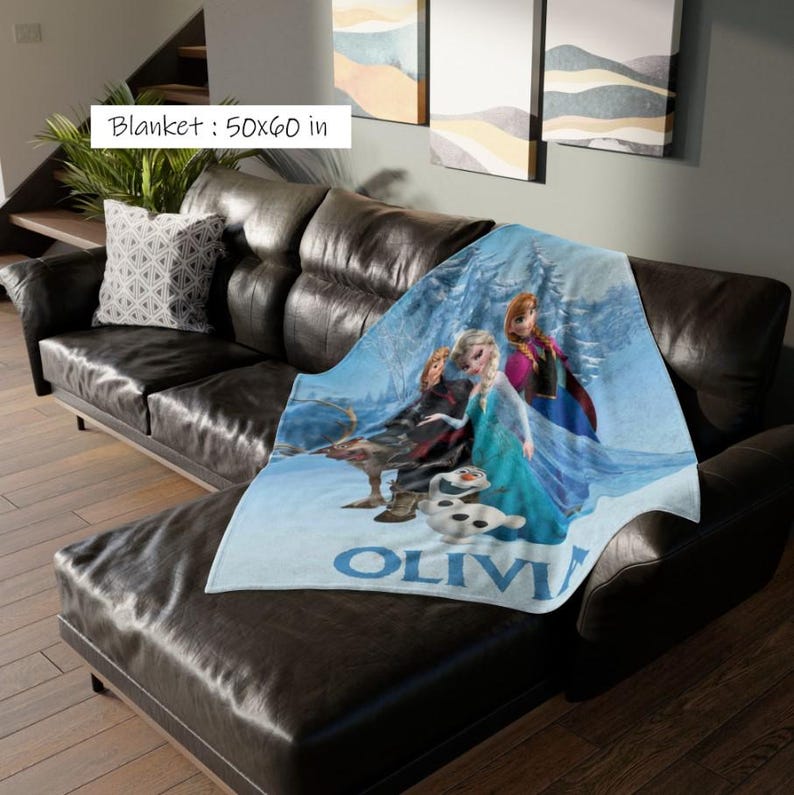 May include: A blue blanket featuring characters from the movie Frozen, with the name "OLIVIA" printed in blue. The blanket measures 50 x 60 inches and is draped over a black leather couch. The blanket's design includes Elsa, Anna, Kristoff, Sven, and Olaf.
