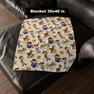 May include: A beige blanket with a repeating pattern of cartoon characters, horses, and stars. The blanket measures 76x102 cm and features the word "Howdy" in a script font. The blanket is folded on a dark leather couch.