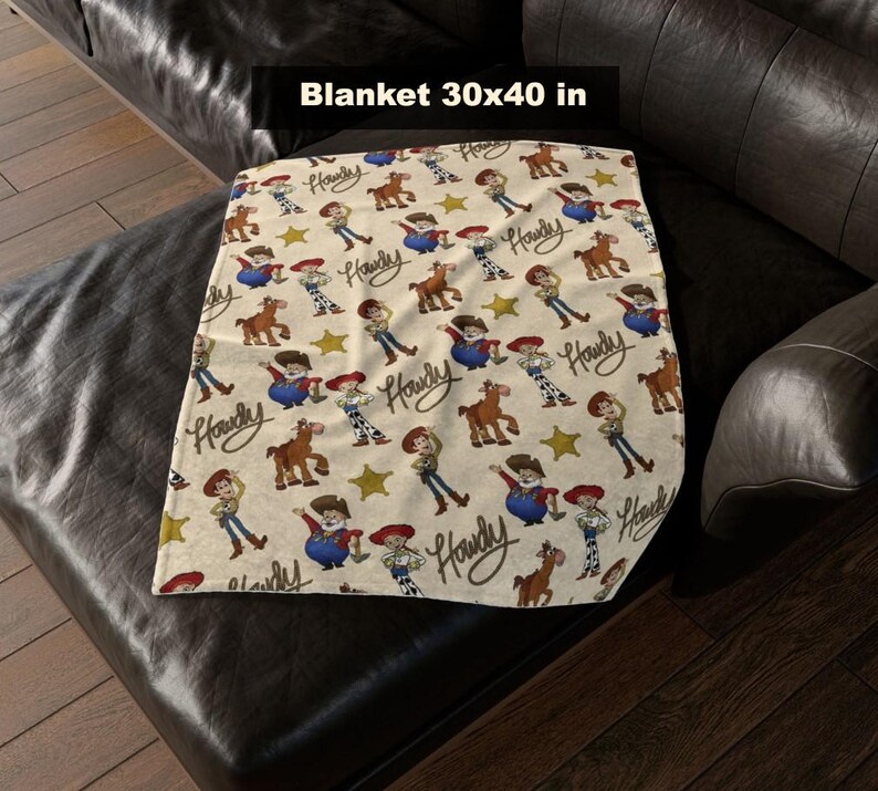 May include: A beige blanket with a repeating pattern of cartoon characters, horses, and stars. The blanket measures 76x102 cm and features the word "Howdy" in a script font. The blanket is folded on a dark leather couch.