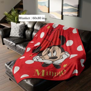 May include: A red blanket with white polka dots and a Minnie Mouse graphic. The blanket, measuring 60x80 inches, features Minnie Mouse with a red bow and the name "Minnie" in gold lettering. The blanket is draped over a black leather couch.
